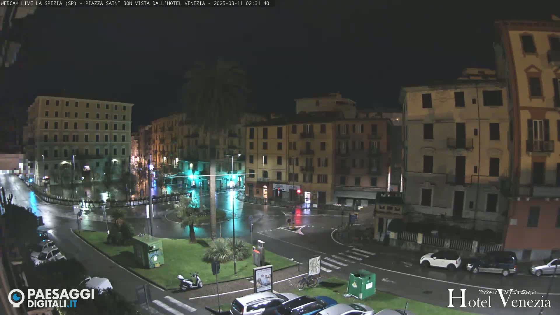 A nighttime view of a rain-slicked town square surrounded by multi-story buildings, with a few cars and a scooter visible.