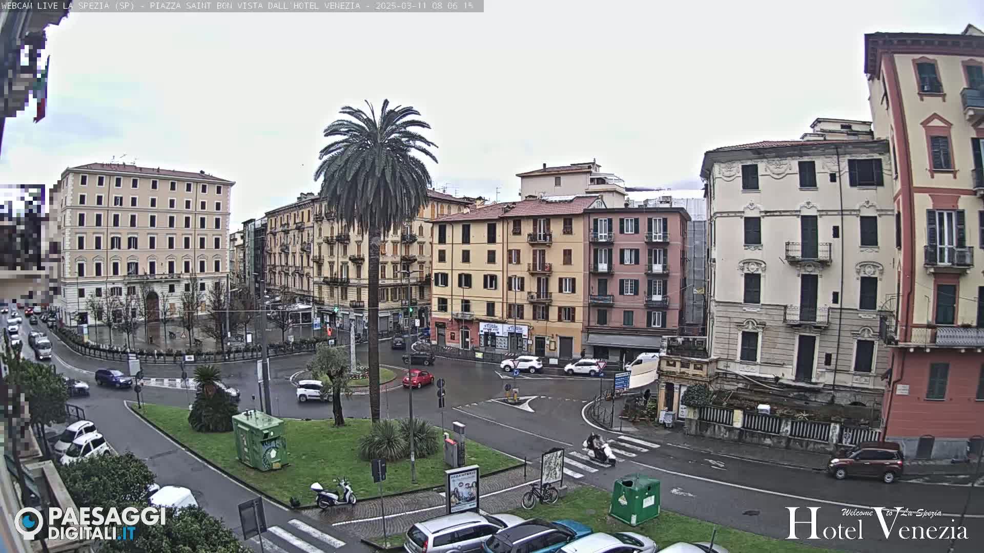 A mostly cloudy day in a European city square with several multi-story buildings surrounding a roundabout with cars and pedestrians; a palm tree sits in the center of the roundabout.