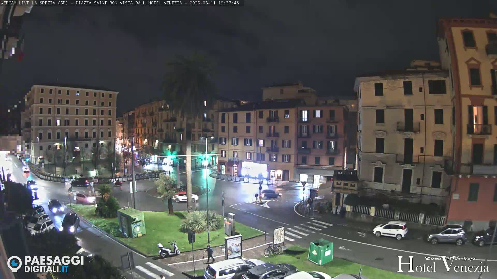 A nighttime view of a city square with several buildings, cars, and a few pedestrians, under a dark, possibly rainy sky.