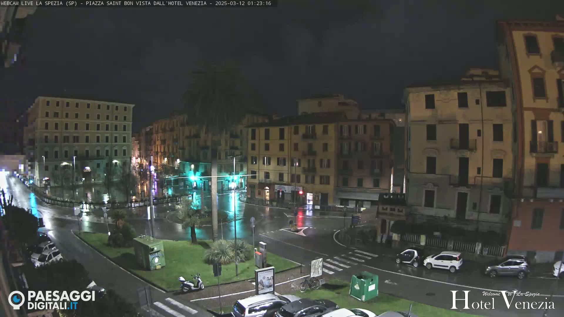 A rain-slicked, nighttime city square is surrounded by multi-story buildings, with a few vehicles and a scooter visible.