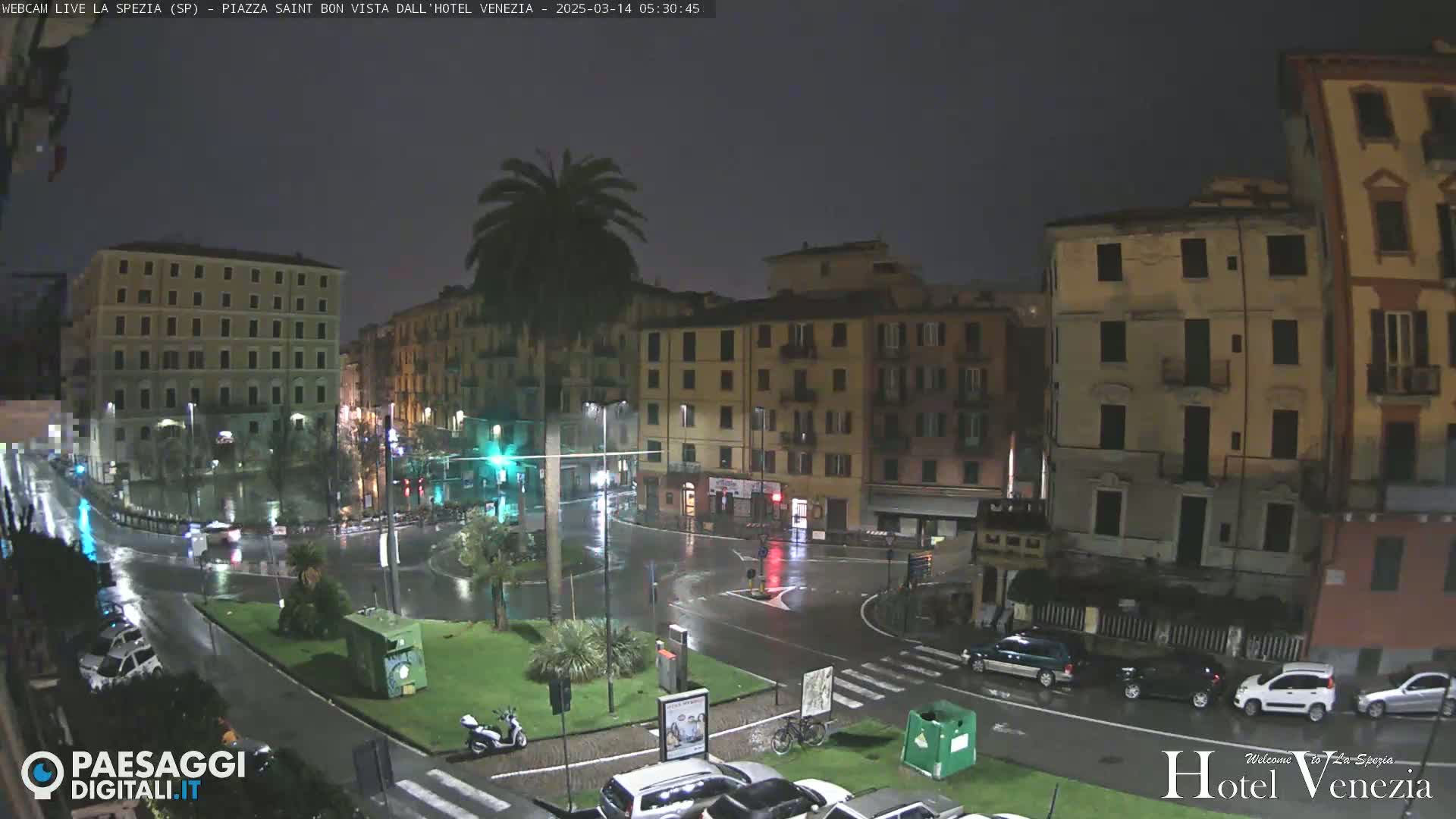 A rain-soaked city square at night, surrounded by multi-story buildings, with a lone palm tree and several cars visible.