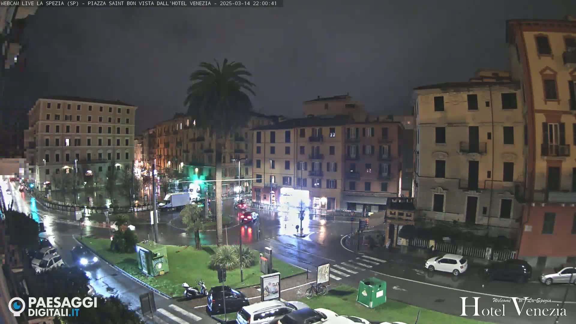 A rain-slicked, nighttime city square surrounded by multi-story buildings, with sparse vehicle traffic and a lone palm tree.