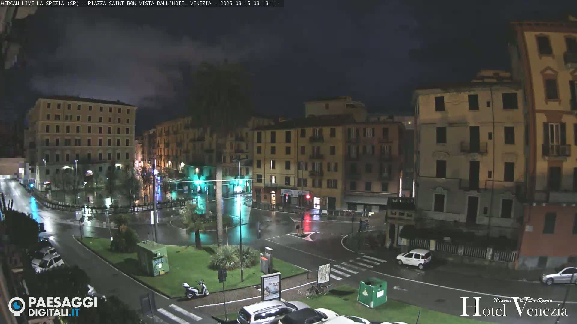 A rain-slicked town square at night, surrounded by multi-story buildings, with a few cars and a scooter visible.