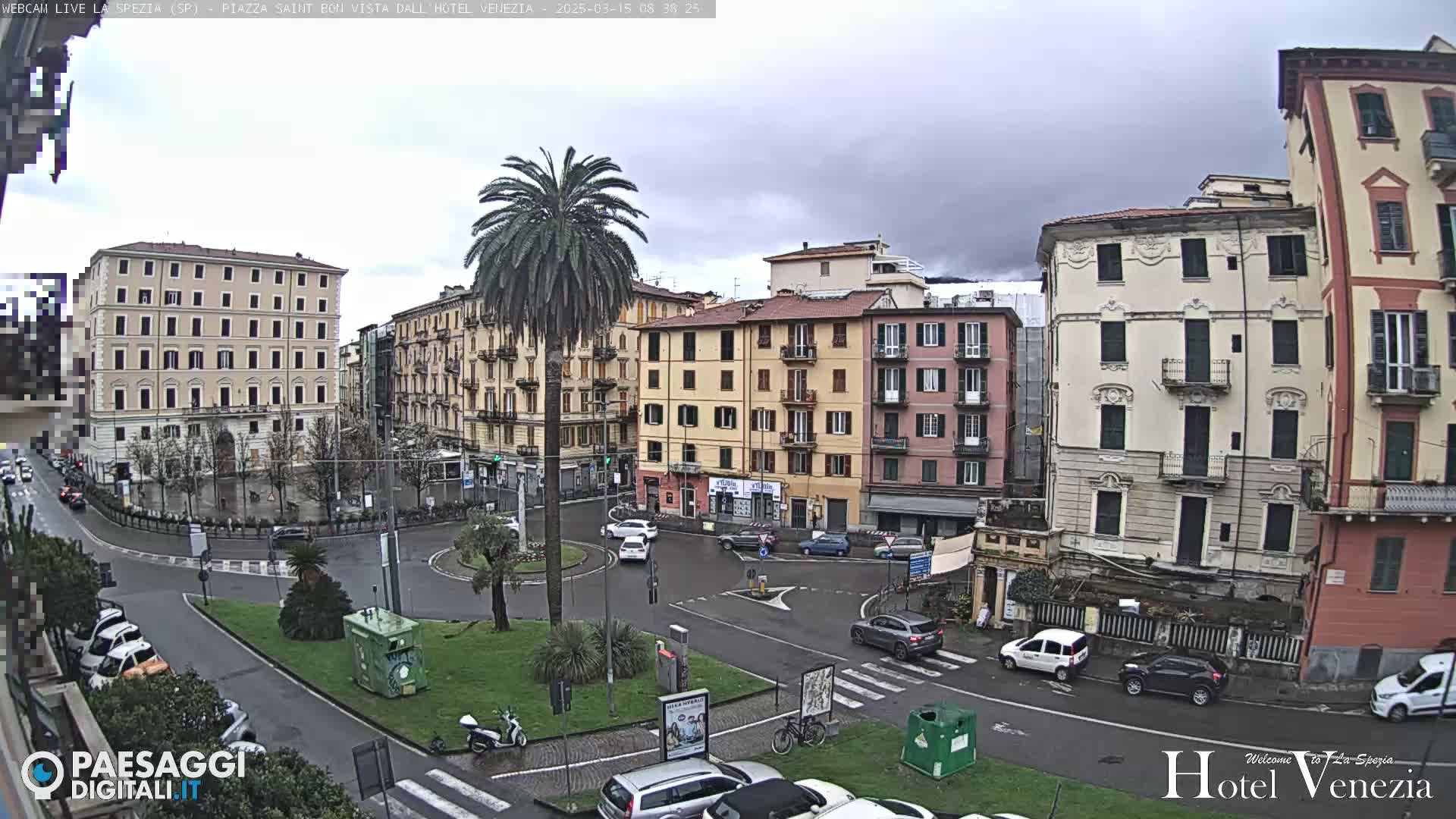 A wet, overcast day in a European city square, featuring several multi-story buildings, a central roundabout with sparse traffic, and a tall palm tree.