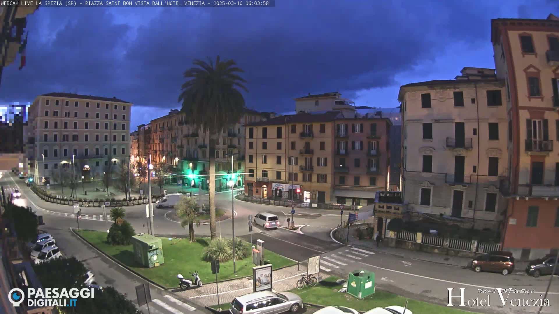 A twilight view of a town square in Italy, under a partly cloudy sky, shows several multi-story buildings surrounding a traffic circle with cars and pedestrians.