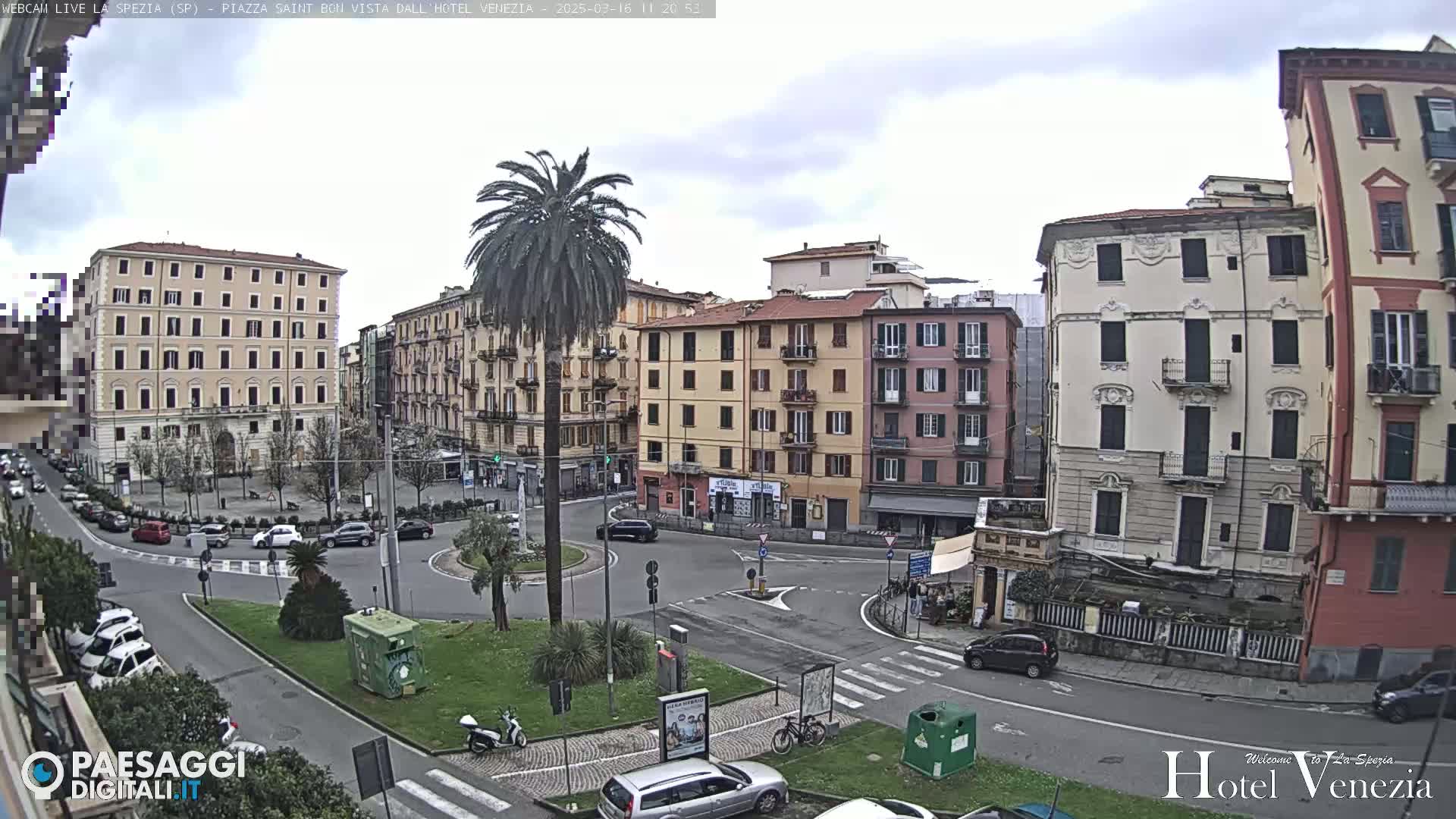 A mostly cloudy day reveals a European city square, with several multi-story buildings surrounding a roundabout where cars and a scooter are visible, and a tall palm tree in the center of the roundabout.