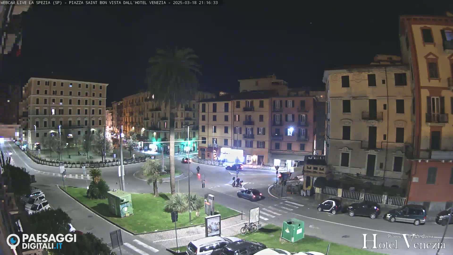 A nighttime view of a town square with several buildings, cars, and a palm tree, under clear skies.