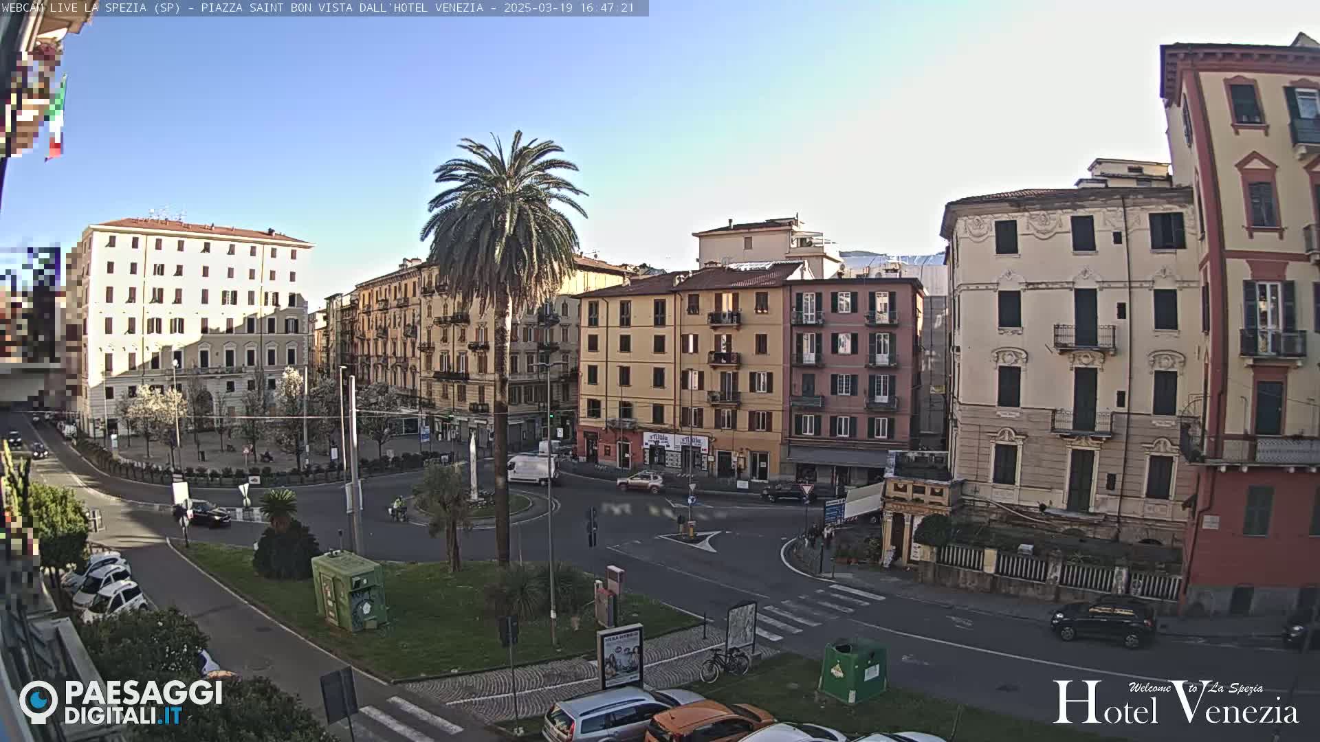 A sunny day reveals a European city square with several multi-story buildings, a central roundabout, and a prominent palm tree.