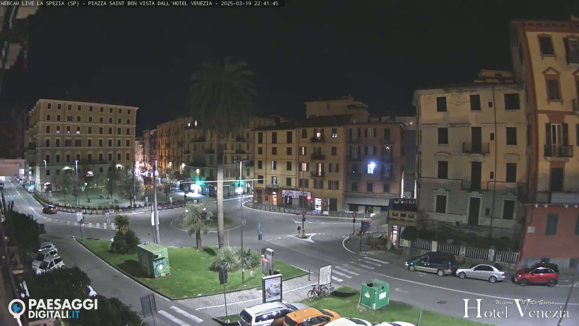 A nighttime view of a plaza surrounded by multi-story buildings in a European city, with cars parked along the streets and a few people visible.