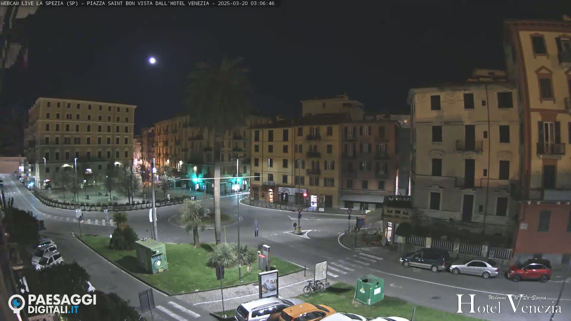 A nighttime, moonlit view of a city square with several multi-story buildings, sparse traffic, and some greenery.