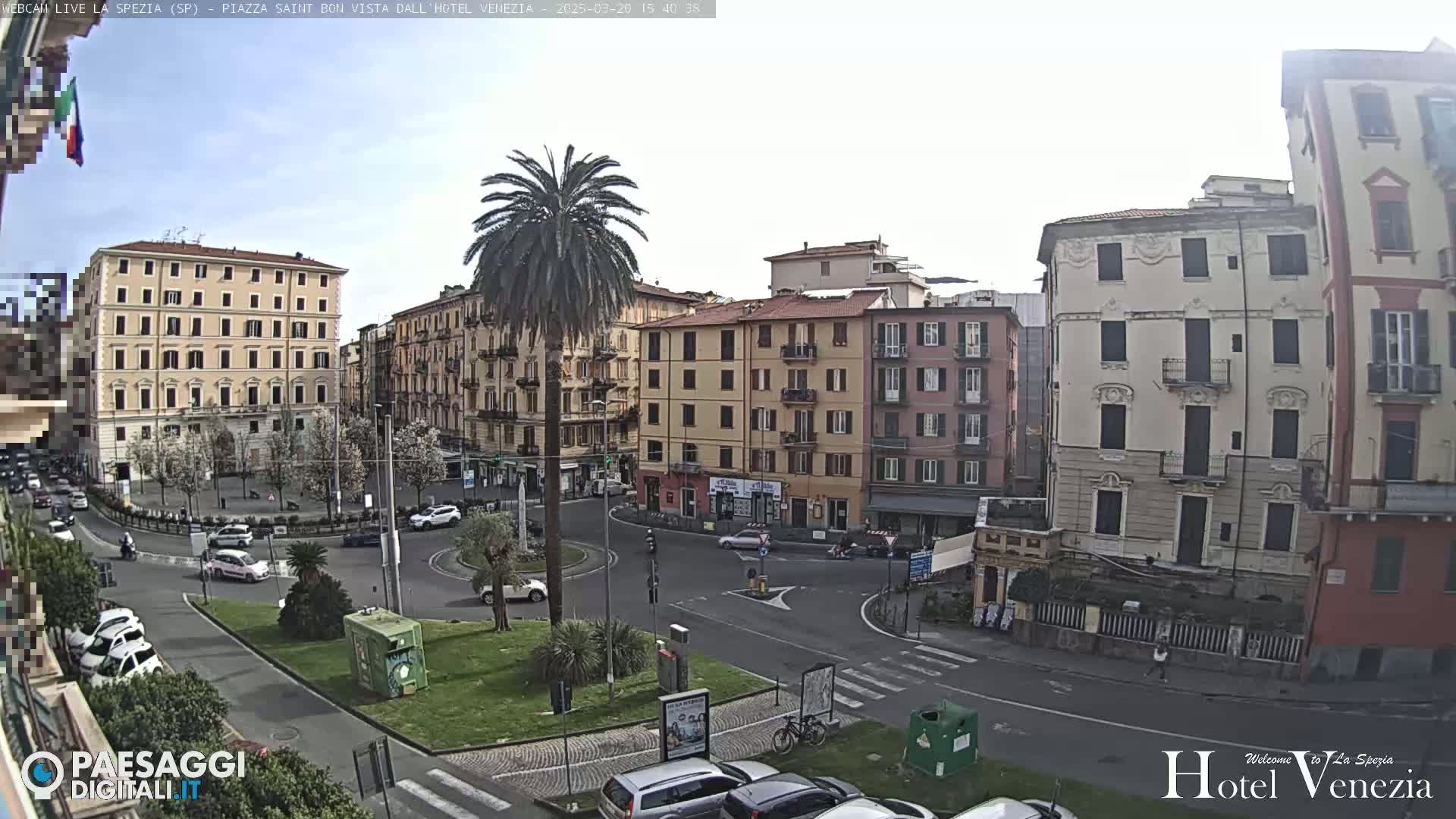 A mostly sunny day reveals a European city square with several multi-story buildings, palm trees, cars, and a roundabout.