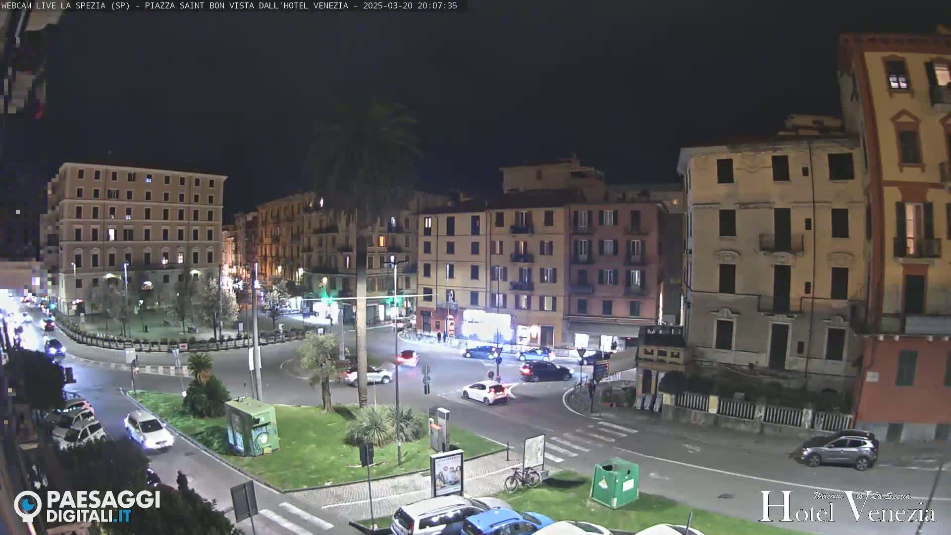 A nighttime view of a city square with several multi-story buildings, cars driving around a roundabout, and a few palm trees.