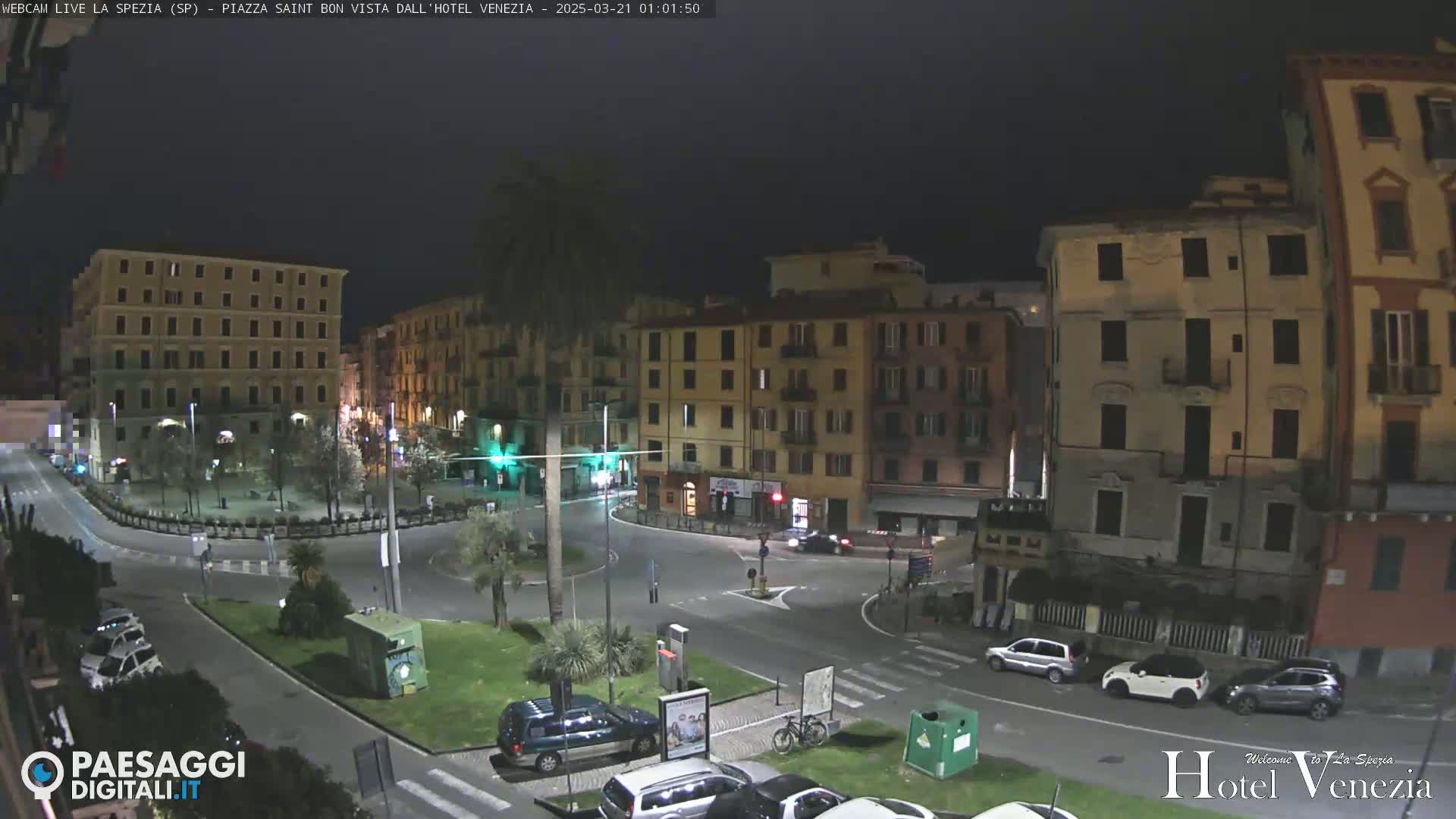 A nighttime view of a mostly empty city square surrounded by multi-story buildings, with a few cars parked and driving around the perimeter.