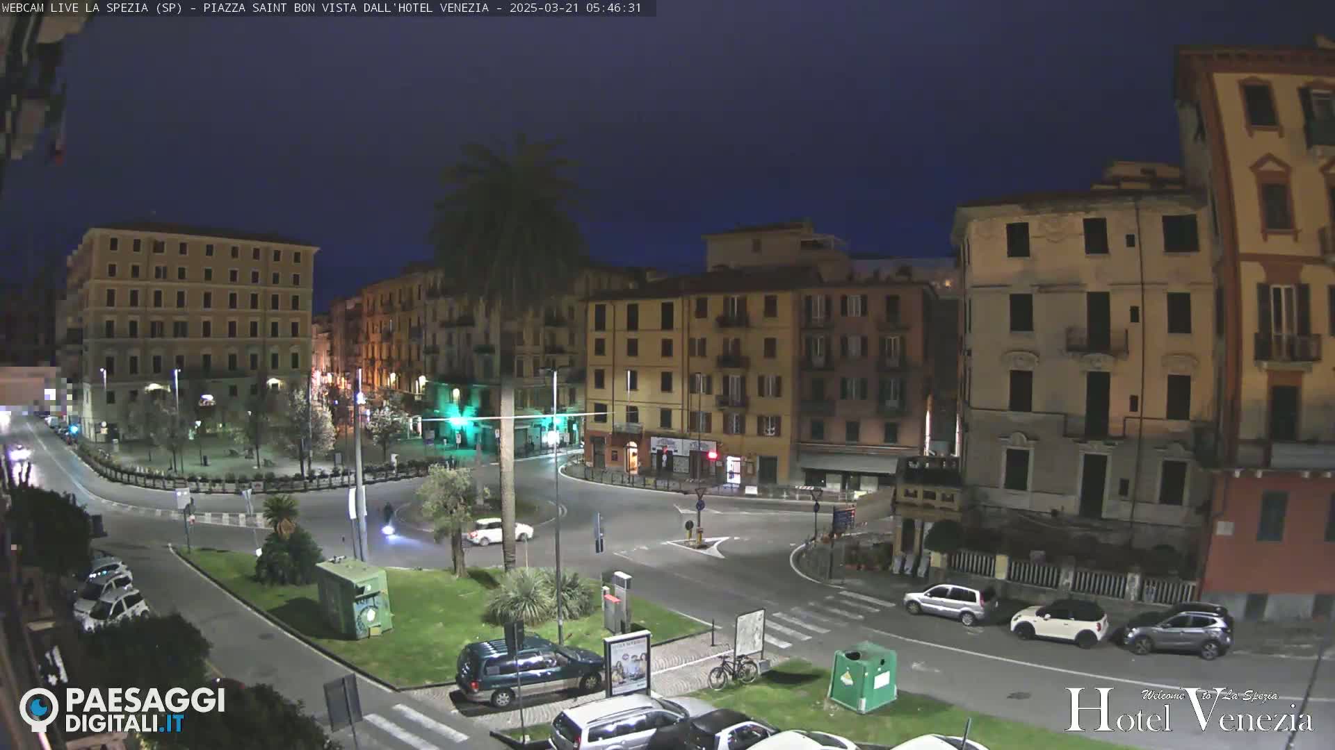 A nighttime view of a mostly empty town square surrounded by multi-story buildings under a dark blue sky.