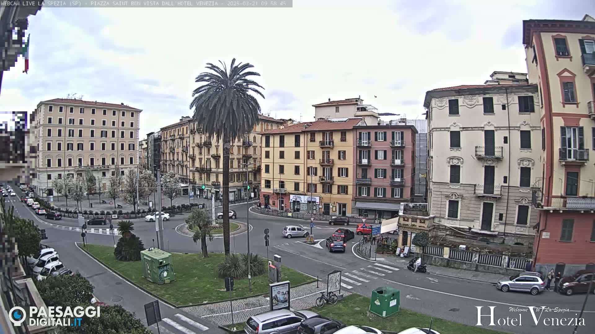 A mostly cloudy day reveals a European-style town square, surrounded by multi-story buildings, with cars parked and driving around a central circular traffic island containing a palm tree.