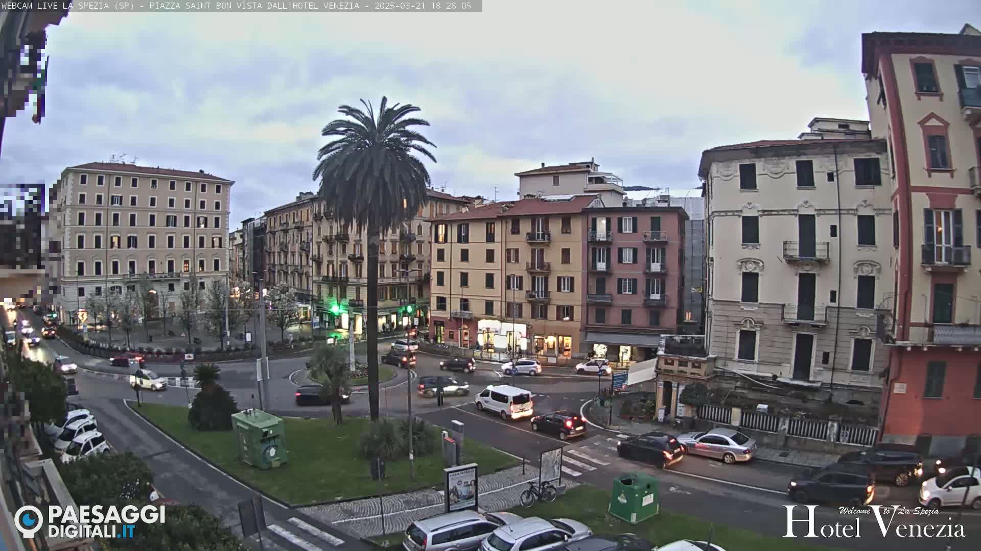 A city square with several multi-story buildings, a palm tree in the center, and cars driving around a roundabout under a cloudy, twilight sky.