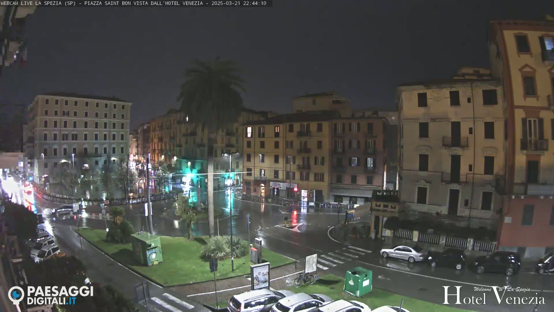 A rain-soaked nighttime city square, surrounded by multi-story buildings, with cars parked and driving on wet streets.