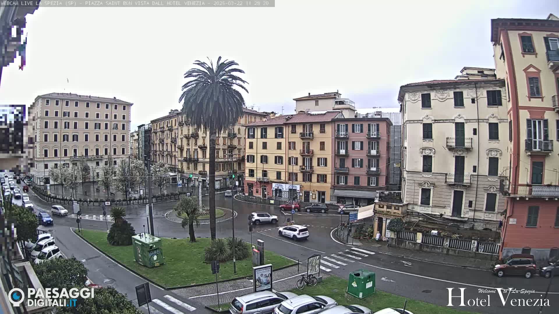 A wet, overcast day reveals a European city square with several multi-story buildings surrounding a roundabout where cars are parked and driving.