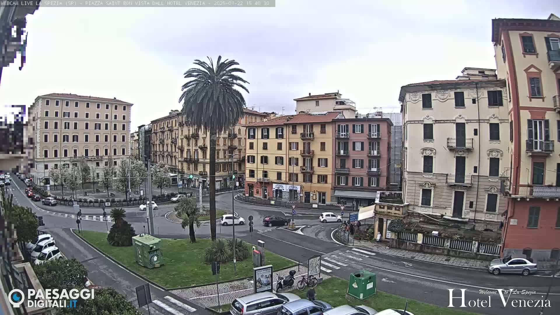 A cloudy day in a European city square, showing several multi-story buildings, a roundabout with cars, and a tall palm tree in the center.