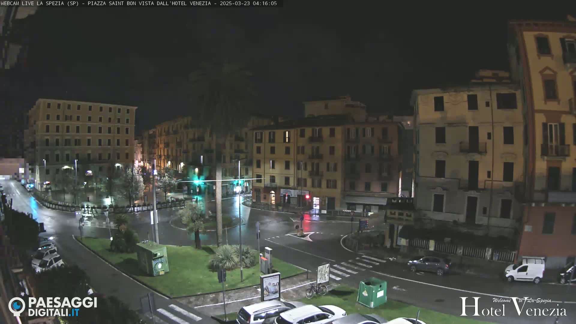 A nighttime view of a rain-slicked, circular plaza surrounded by multi-story buildings, with a few cars and a bicycle visible.