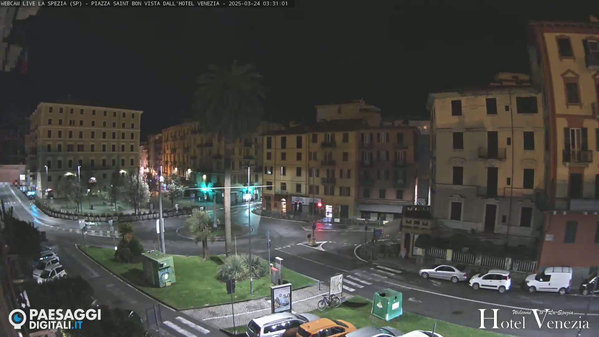 A nighttime, wet view of a town square surrounded by multi-story buildings and a few parked cars.