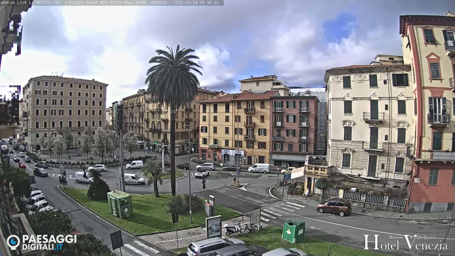 A partly cloudy day reveals a city square with several multi-story buildings, a central roundabout with cars and a palm tree, and surrounding green landscaping.