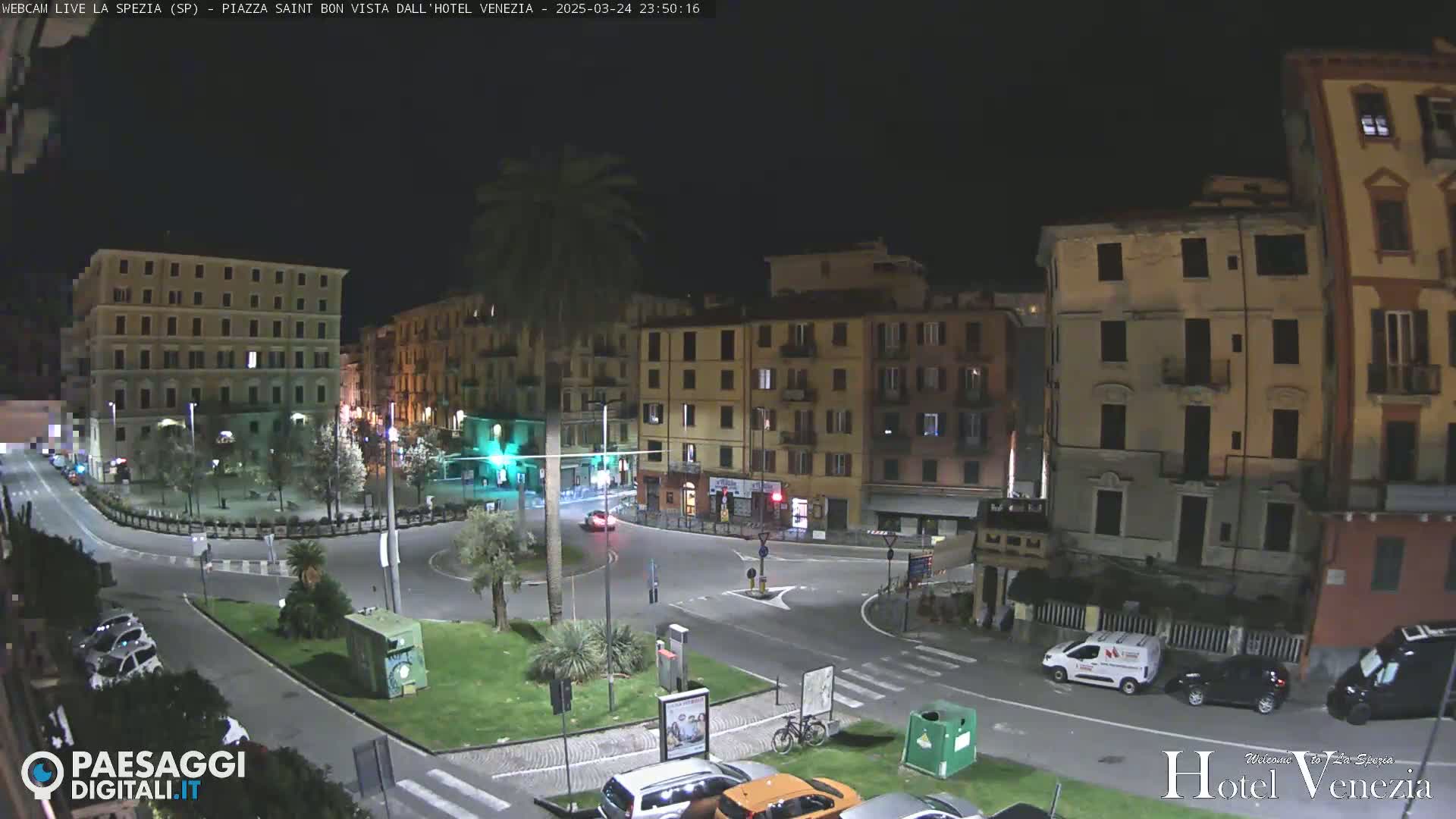 A nighttime view of a plaza in a city with several multi-story buildings surrounding a small roundabout, where a few cars and a van are parked.