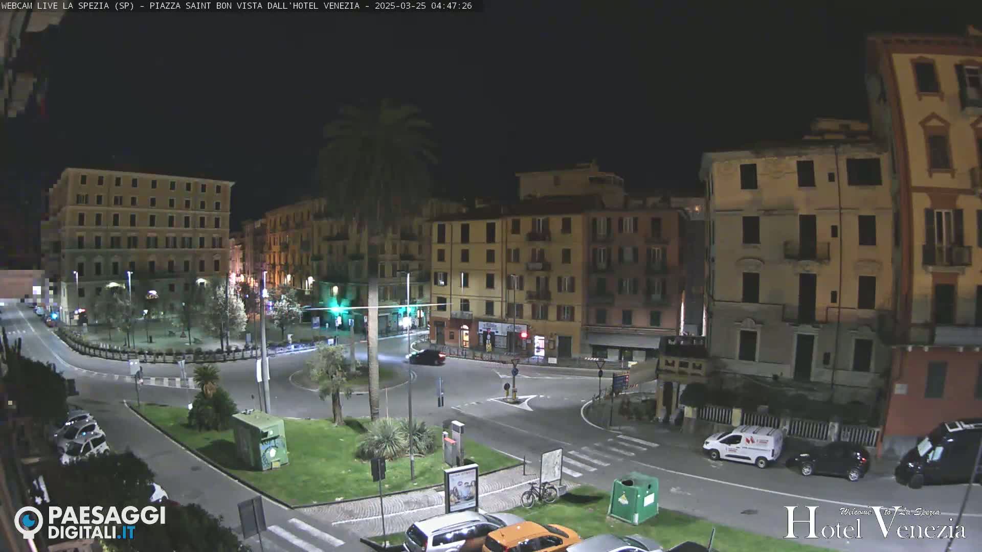 A nighttime view of a plaza in a European city with several multi-story buildings surrounding a circular road, and cars parked along the street.