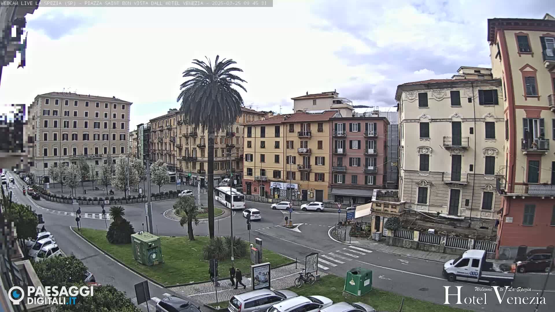 A mostly cloudy day reveals a circular traffic intersection surrounded by multi-story buildings in a European-style city square, with a tall palm tree in the center of the square.
