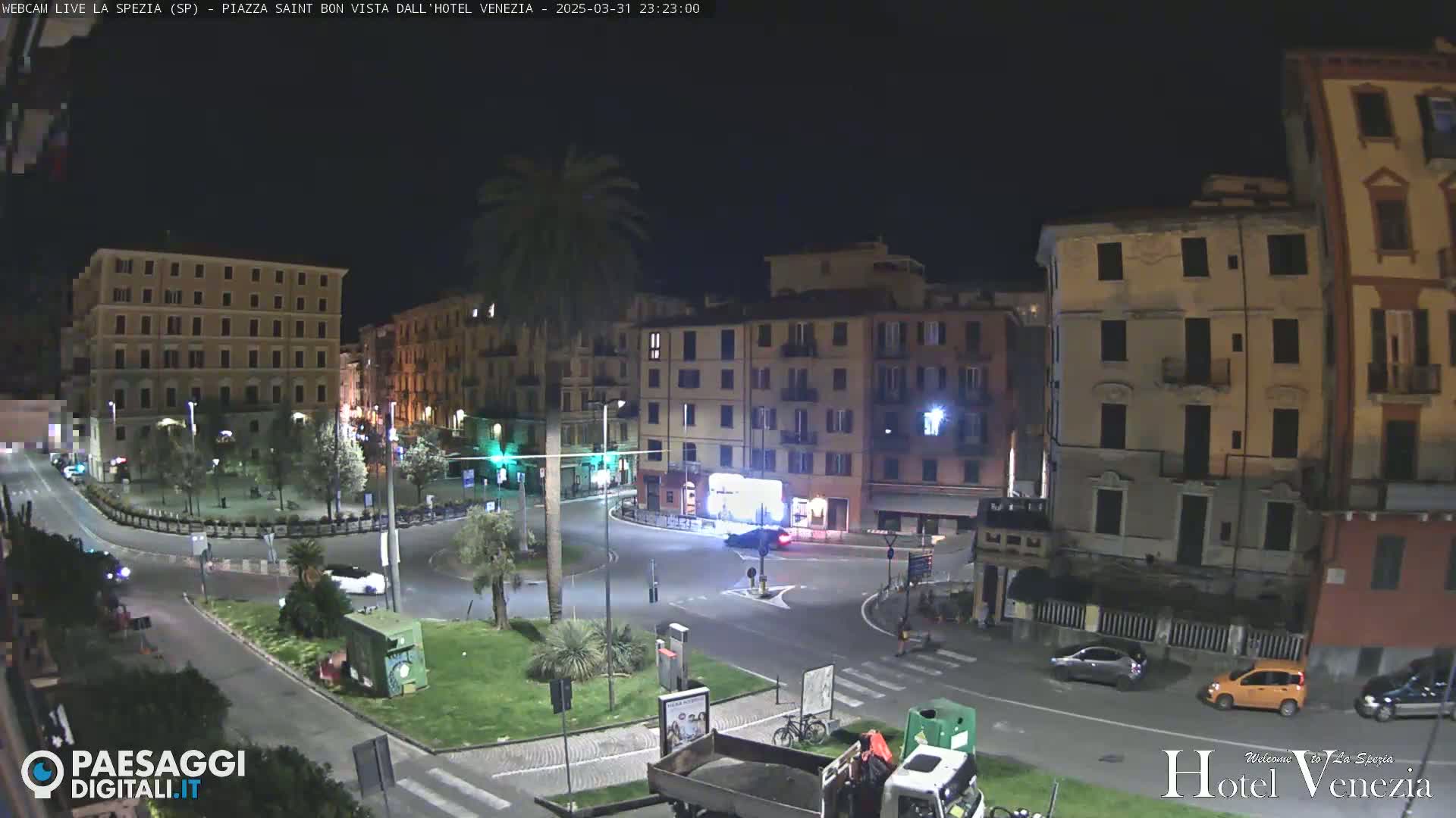 A nighttime view of a mostly empty town square with several multi-story buildings surrounding it, a few cars, and a lone palm tree.