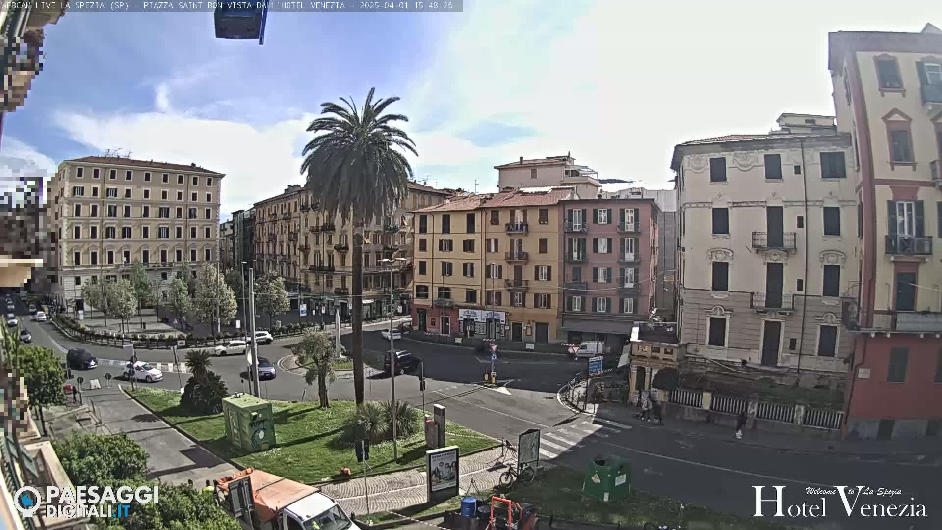A sunny day in a European city square, with several multi-story buildings surrounding a roundabout with a palm tree in the center.