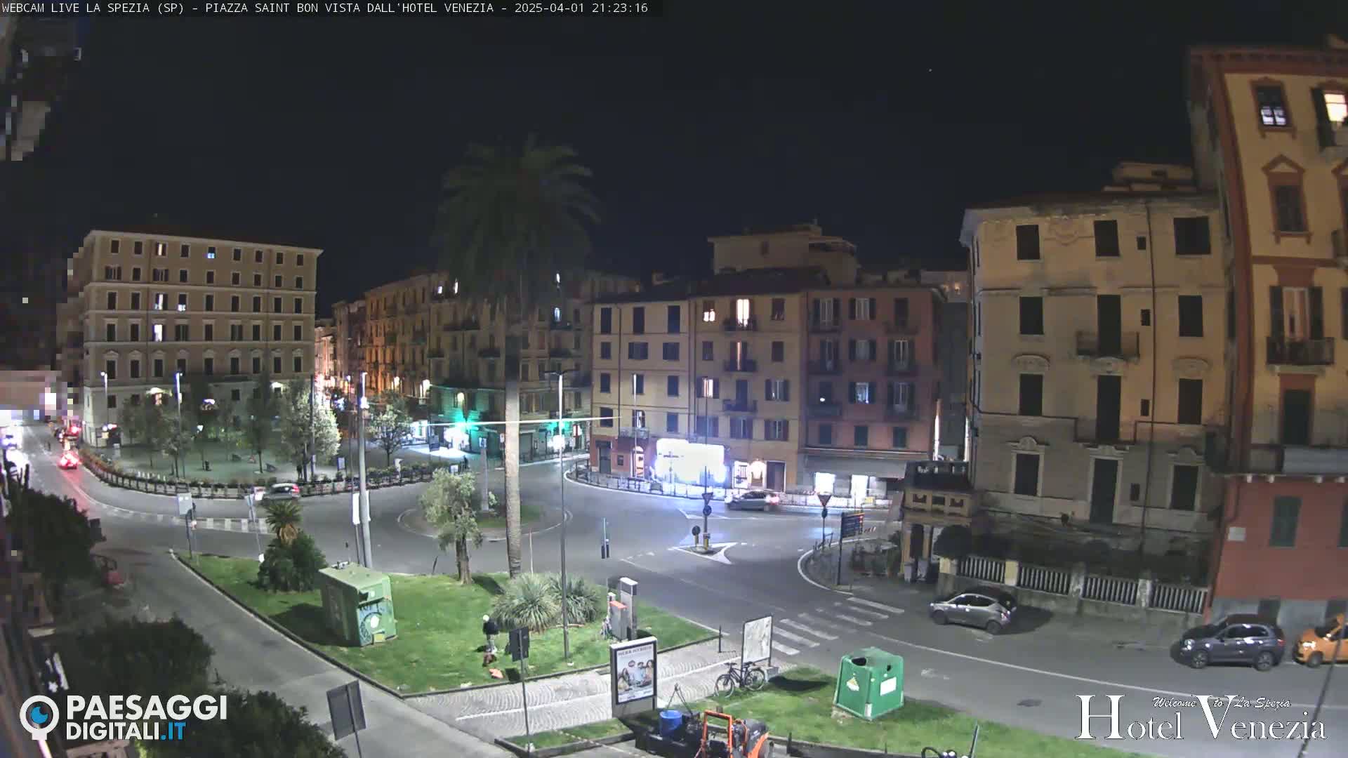 A nighttime, high-angle view of a city square with several buildings surrounding it, cars parked along the streets, and some people walking around.