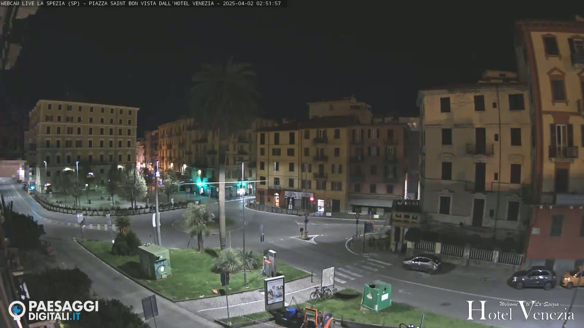 A nighttime view of a quiet town square surrounded by multi-story buildings, with a few cars parked along the roadside and some greenery in the center.