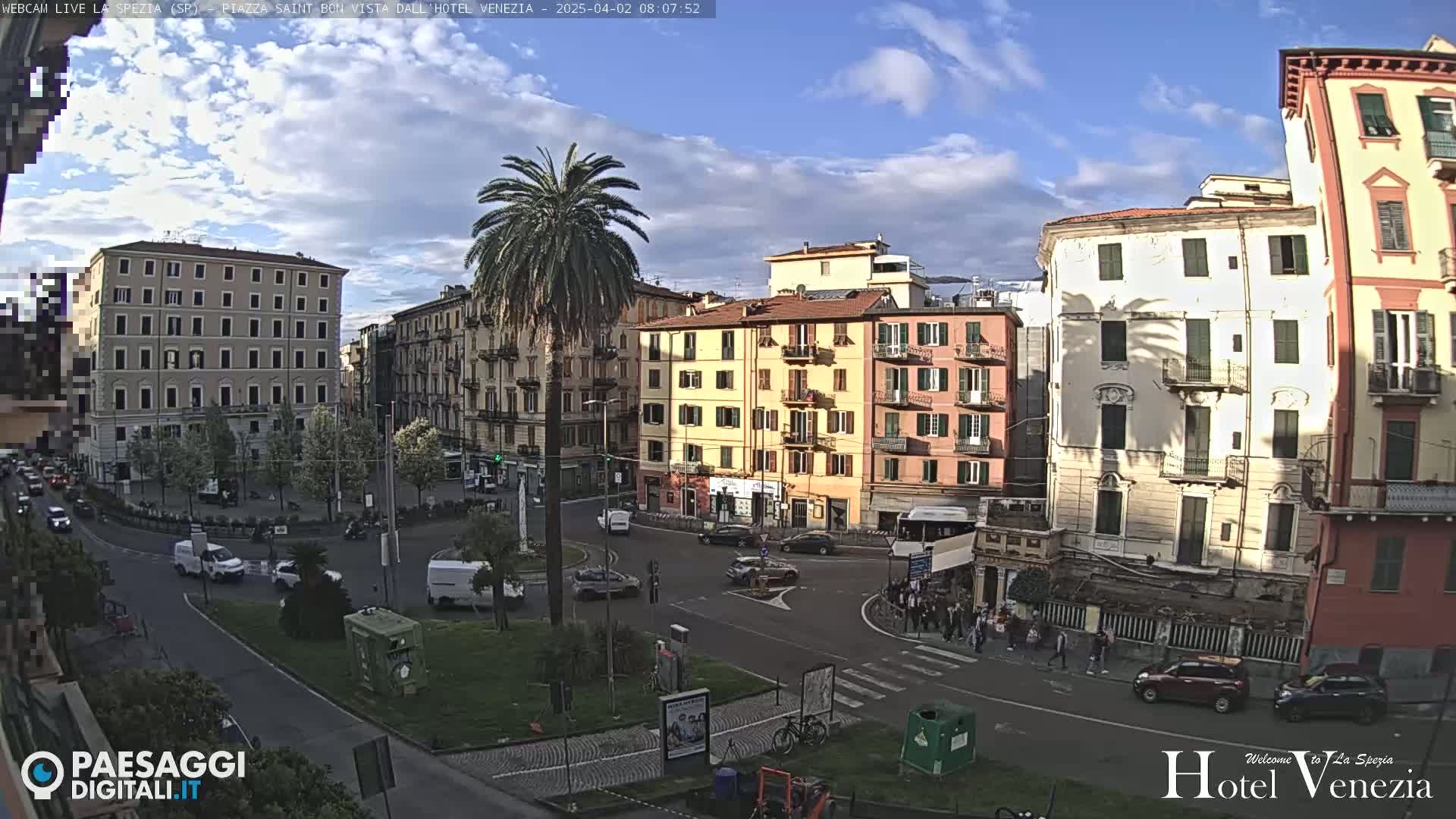 A sunny day reveals a city square surrounded by multi-story buildings, with cars and pedestrians visible, and a palm tree in the center.