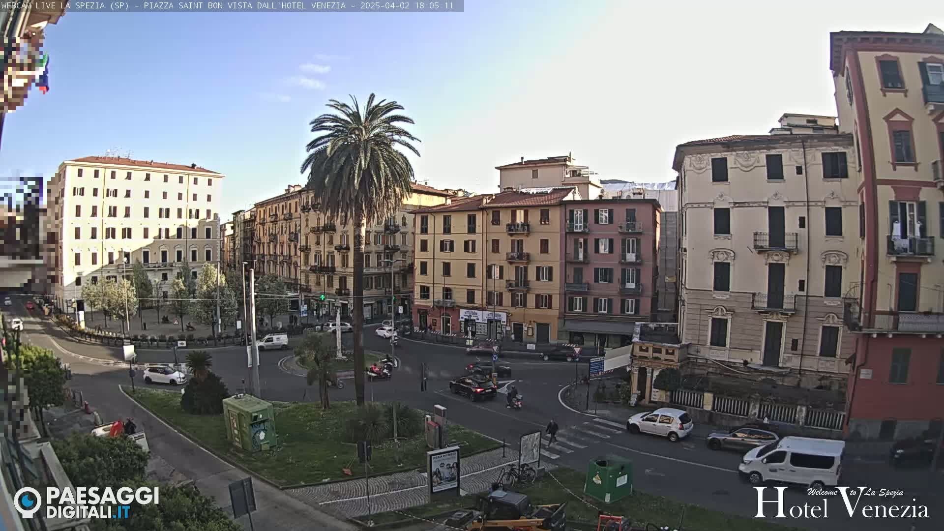 A sunny day reveals a city square with several multi-story buildings, a palm tree in the center, and cars driving around a roundabout.