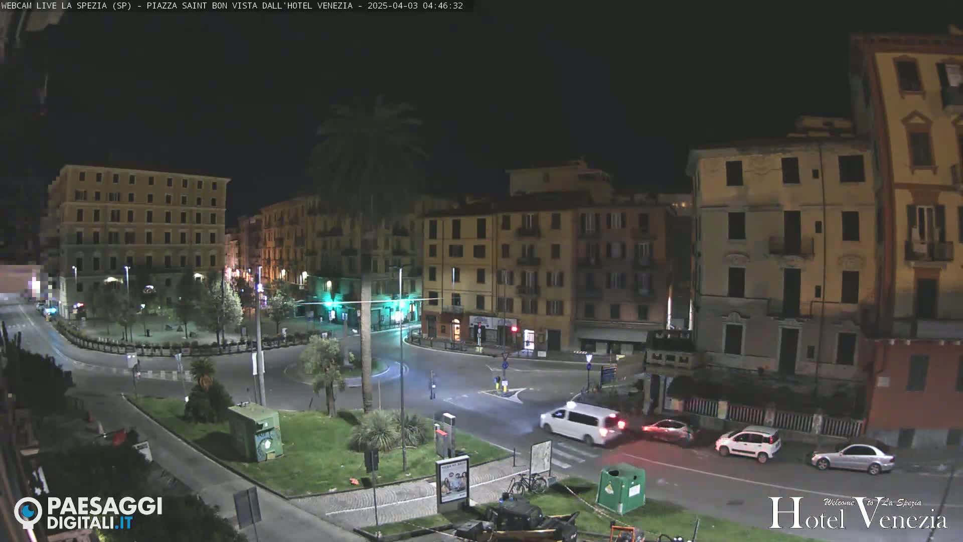 A nighttime view of a city square with several multi-story buildings, sparse traffic, and a few palm trees, under clear night skies.