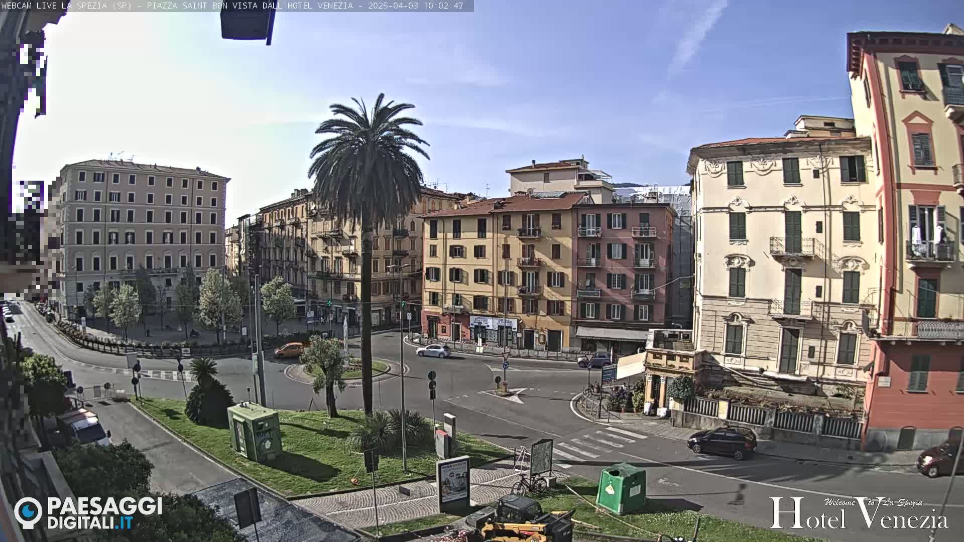 A sunny day in a European city square, showing several multi-story buildings and a roundabout with sparse traffic.