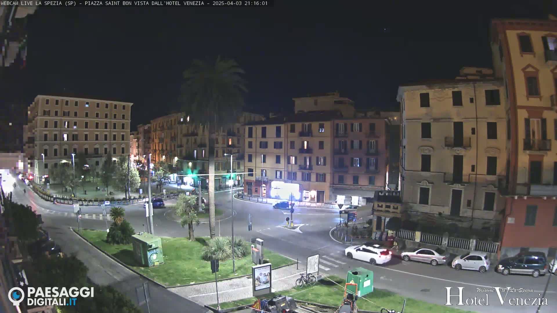 A nighttime view of a city square with several multi-story buildings surrounding a traffic circle, where a few cars are parked and driving.
