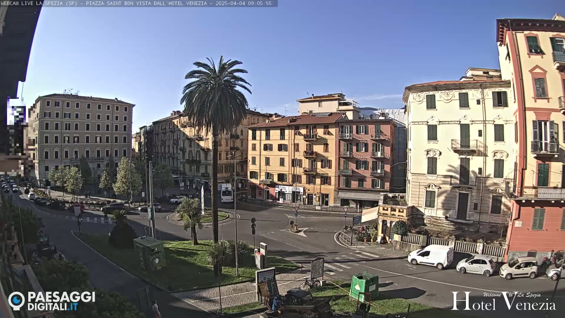 A sunny day reveals a European city square with several multi-story buildings, a palm tree, and a few vehicles parked and driving around a roundabout.
