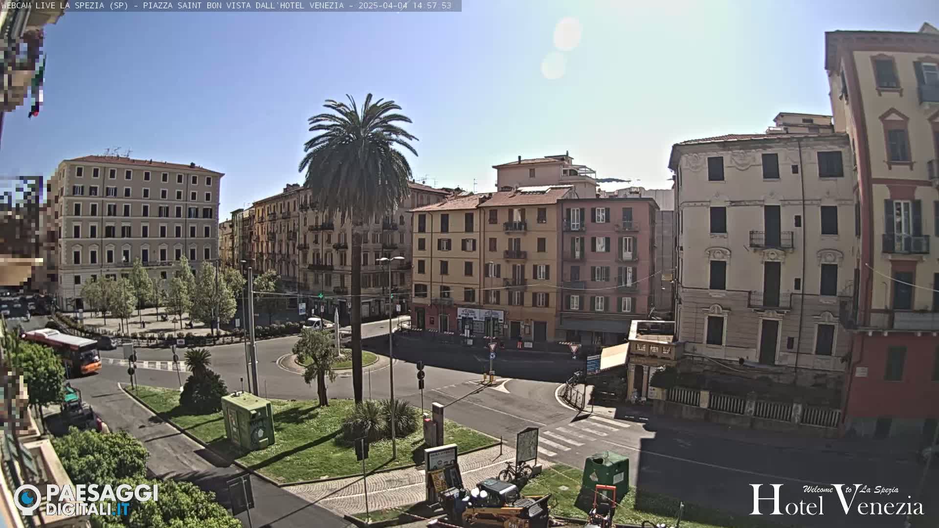 A sunny day in a European city square, showing several multi-story buildings surrounding a roundabout with a palm tree in the center, and some vehicles parked nearby.