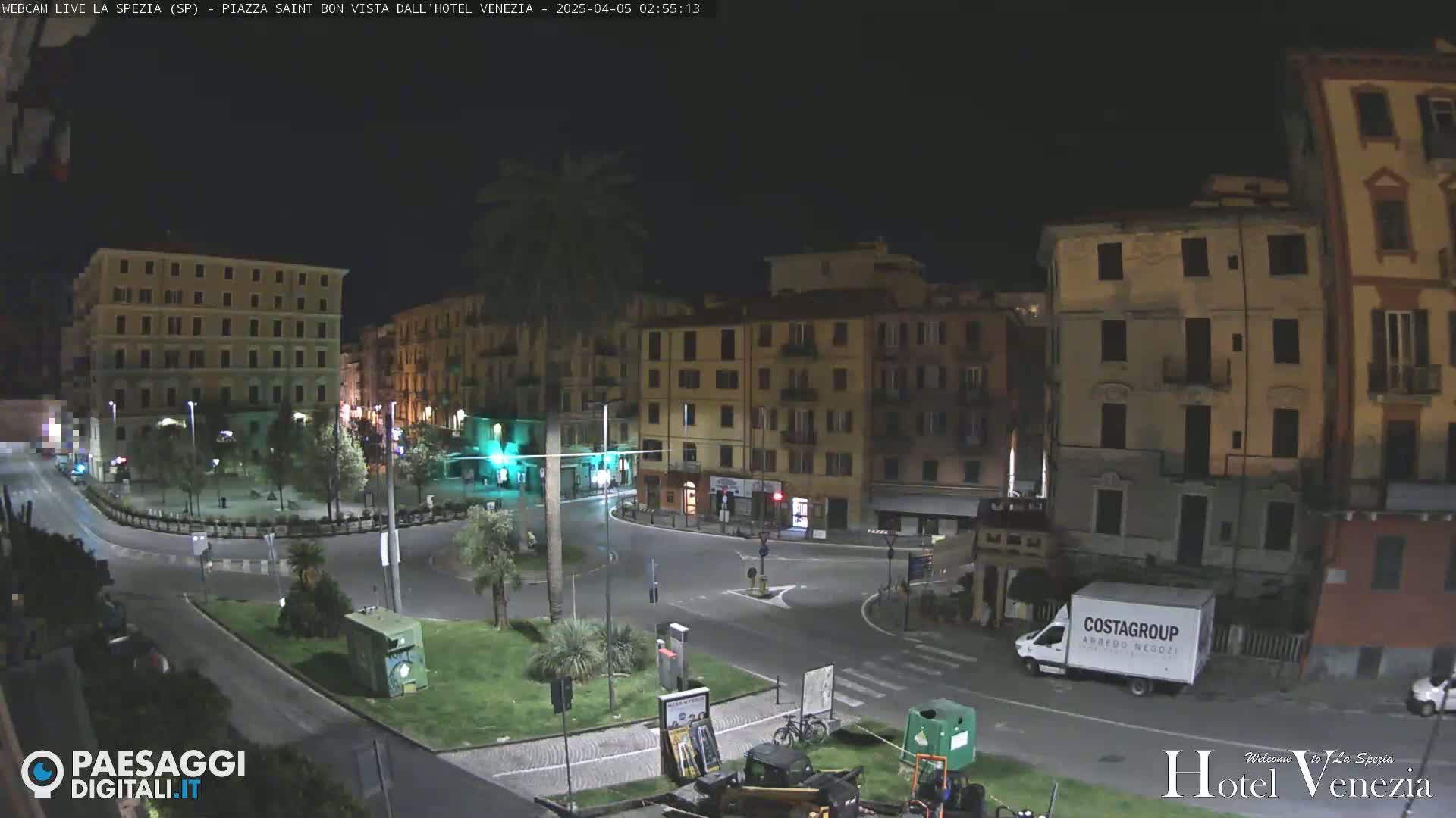 A nighttime view of a mostly empty town square surrounded by multi-story buildings, with a few vehicles and some landscaping visible.