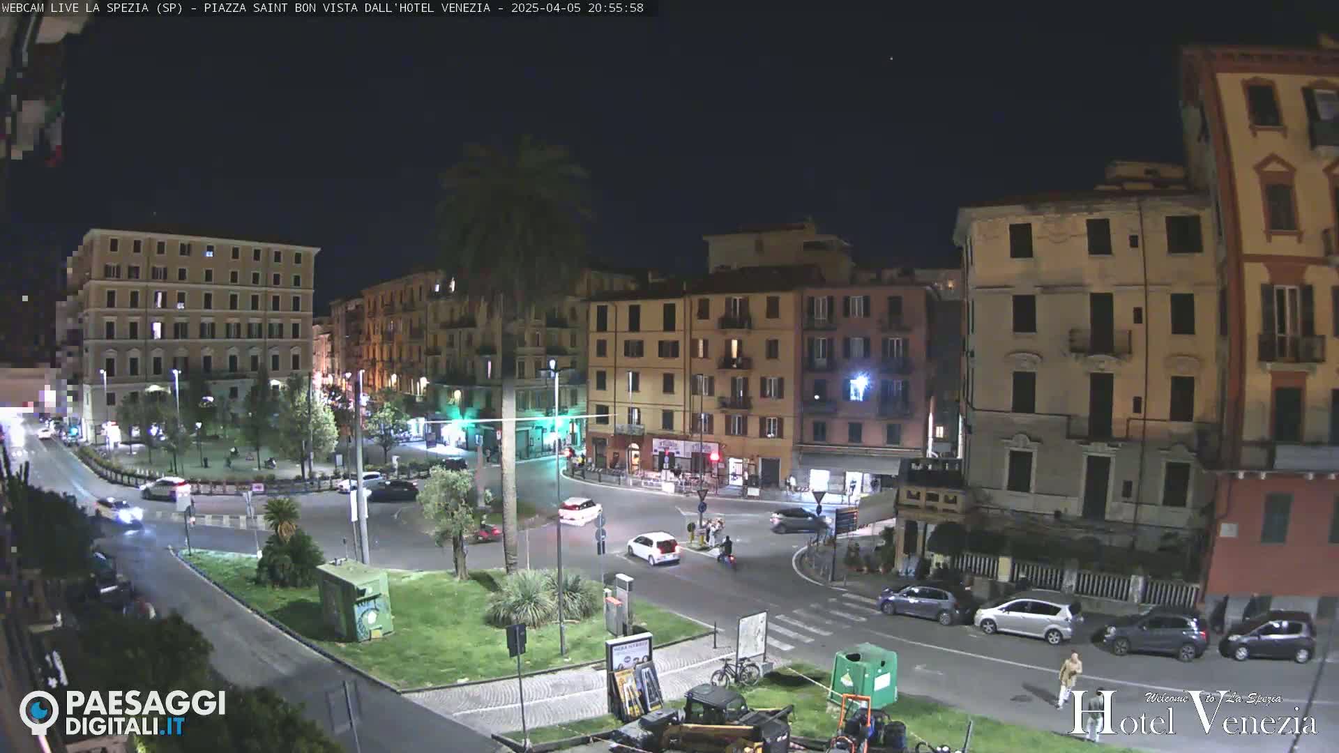 A nighttime, clear-weather view of a city square with several buildings surrounding it, some cars are driving on the roadways, and there are people walking near a small plaza.