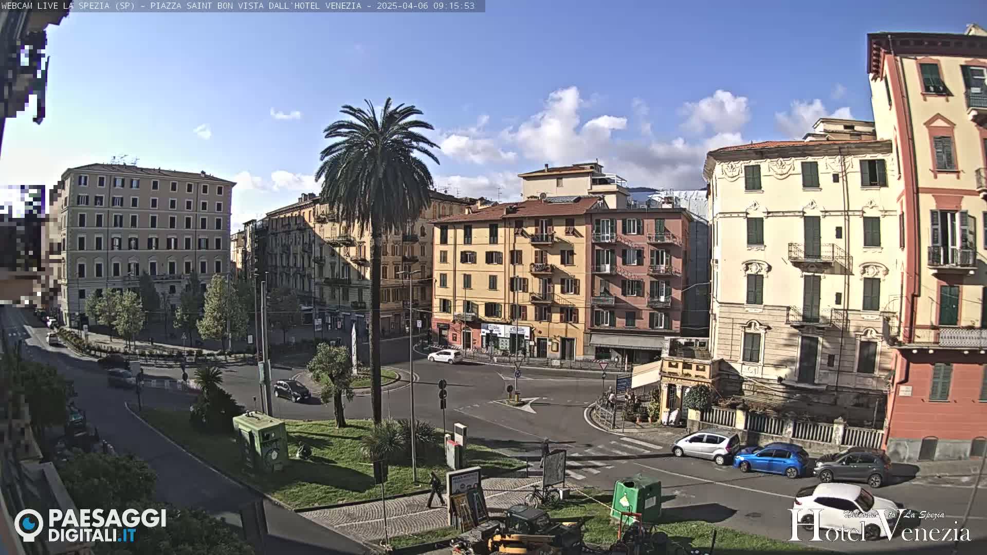 A sunny day in a European city square, with several multi-story buildings surrounding a roundabout with cars and a palm tree in the center.