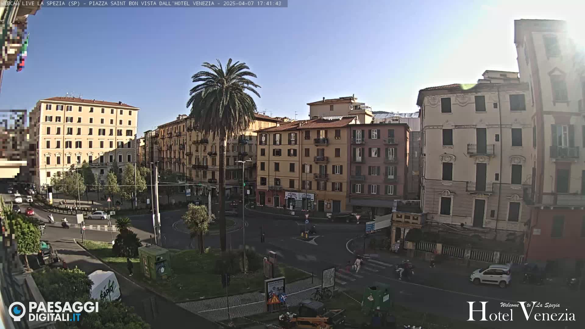 A sunny day reveals a European city square, with several multi-story buildings surrounding a central roundabout where cars and people are moving.  A tall palm tree stands in the center of the square.
