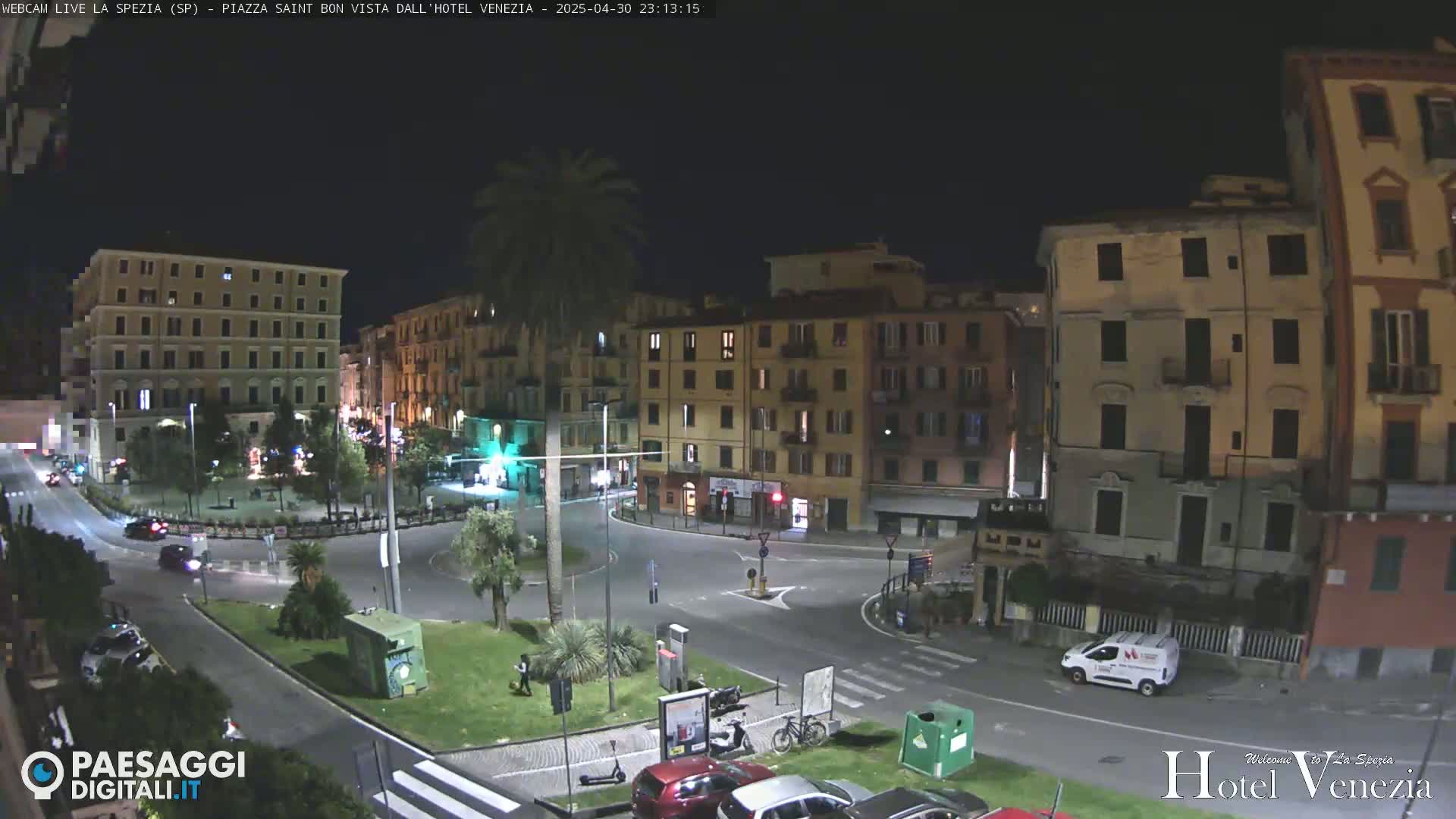 A nighttime, city-center roundabout scene with several multi-story buildings, palm trees, and sparse vehicle traffic.