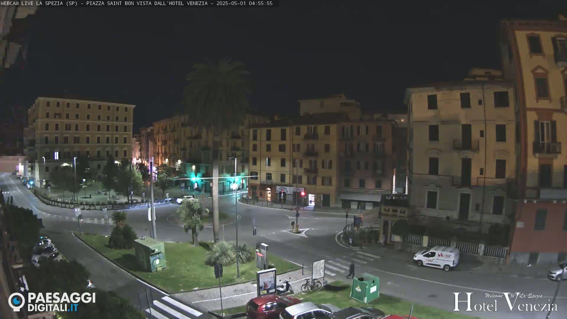 A nighttime view of a mostly empty town square surrounded by multi-story buildings, with a few cars and pedestrians visible.