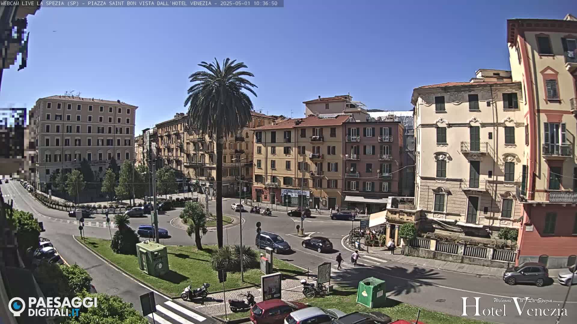A sunny day in a European city square shows several multi-story buildings, palm trees, cars, and pedestrians, with a roundabout at the center.