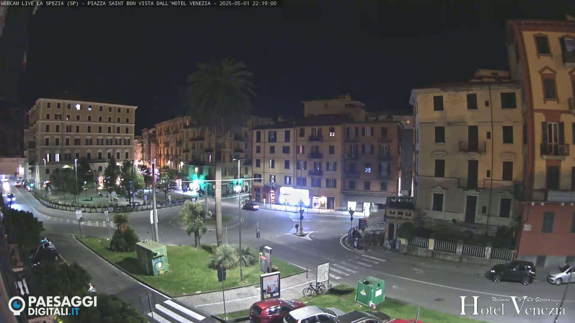 A nighttime view of a city square with several multi-story buildings surrounding a roundabout, and a few cars visible.