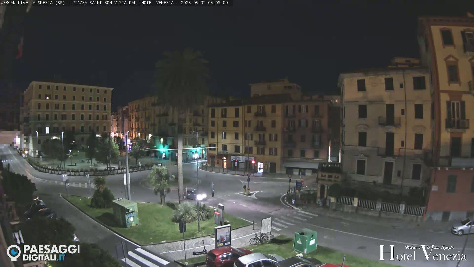 A nighttime view of a city square with several multi-story buildings surrounding a traffic circle, with a few cars and a bicycle visible.