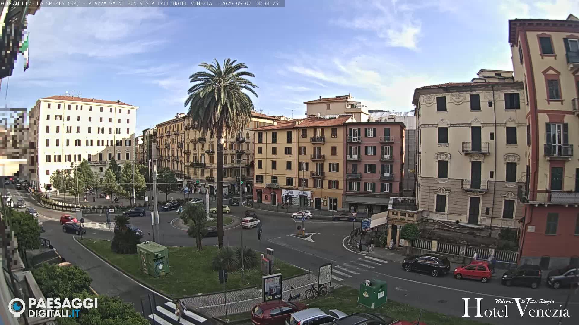 A sunny cityscape with several multi-story buildings surrounding a small plaza, where cars are driving and a palm tree stands in the center.